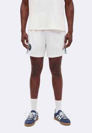 Homme portant un short de sport blanc avec des écussons logo, des chaussettes blanches et des baskets Adidas bleues, debout devant un fond uni.