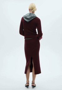 Bordeauxfarbenes, geripptes Strickpullover mit grauem Kapuzen, kombiniert mit einem passenden Rock mit Schlitz hinten. Das Modell steht mit dem Rücken zur Betrachtenden.