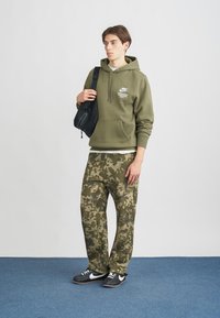 Noor mees, kes kannab oliivroheline Nike huppi, kamuflaažikanga pükse, musti Nike spordijalatsid ja kannab musta õlakotti, seisab sinisel vaibal valge seina ees.