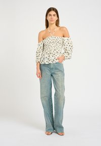 Giovane donna in piedi con una blusa floreale bianca senza spalline con bretelle halter e jeans a vita alta e gamba larga decorati con piccoli rivetti.