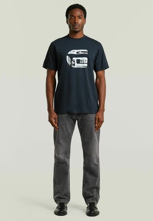 Mand stående vendt fremad iført en sort grafisk t-shirt, grå jeans og sorte sko mod en ensfarvet lys baggrund.