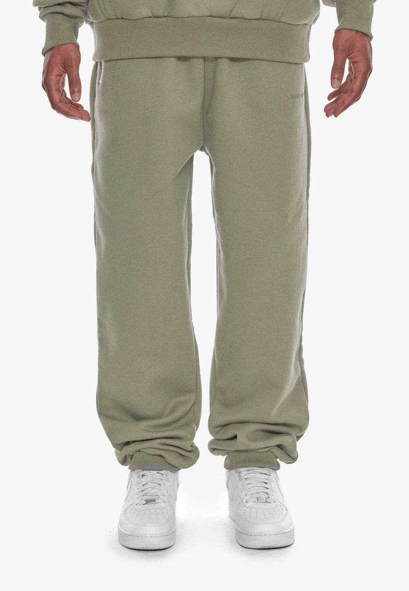 Olivengrønne sweatpants i blødt stof, der har en løs pasform med elastiske manchetter ved anklerne og et subtilt logo på siden.
