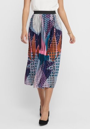 Femme portant une jupe midi plissée aux motifs géométriques et à pois en rose, bleu, orange et noir, associée à des talons noirs.