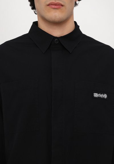 Camisa negra de botones, hecha de algodón, con un cuello estructurado, dos bolsillos en el pecho y un bolsillo izquierdo que presenta un logotipo bordado.