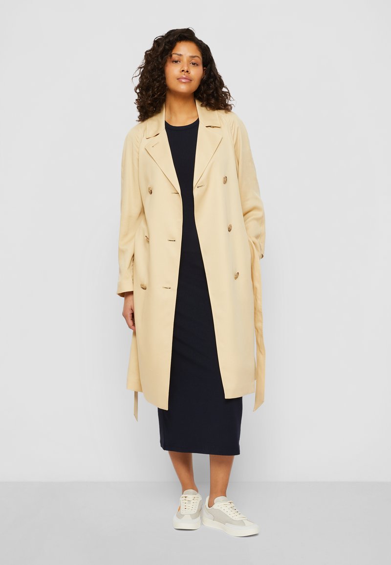 Trench coat beige doppiopetto con una consistenza morbida, cinturini in vita regolabili e bottoni grandi, abbinato a un vestito blu navy a righe e sneakers bianche.