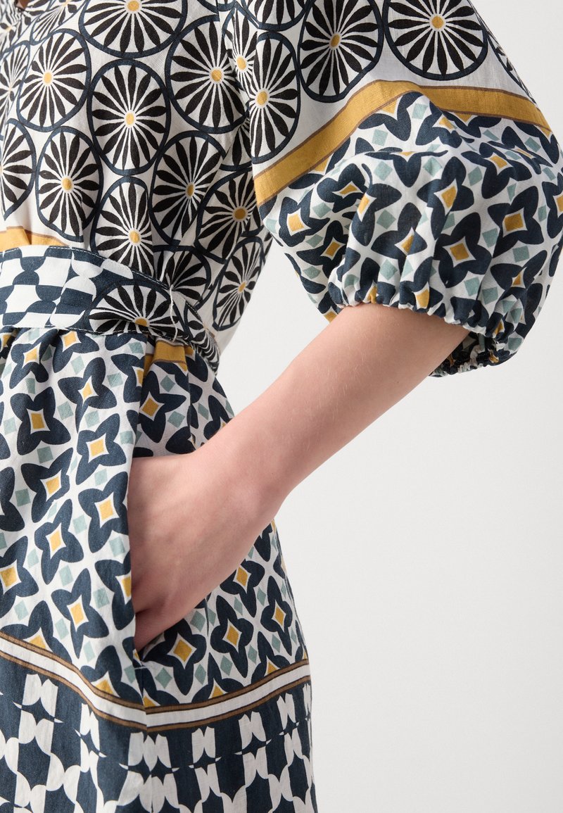 Gemustertes Kleid mit kreisförmigen und geometrischen Designs in Schwarz, Weiß, Marineblau und Gelb. Elastische Ärmel, Taillenkordel und seitliche Tasche sind sichtbar.