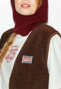 Gilet en polaire marron avec un col relevé en bordeaux. La chemise blanche en dessous affiche un texte imprimé rouge et une étiquette bleue sur le gilet.
