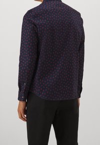 Man som bär en marinblå långärmad skjorta med ett litet rött lövmönster, kombinerad med svarta byxor, sedd bakifrån.