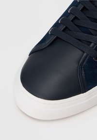 Baskets en cuir et daim bleu marine avec un embout arrondi, une surface texturée et une semelle blanche en caoutchouc. Elles sont dotées de lacets plats et d'œillets en métal.