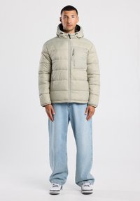 Chaqueta acolchada en gris claro, con costuras horizontales, cierre de cremallera, bolsillo frontal y capucha fija. Llevado con pantalones vaqueros azules holgados.