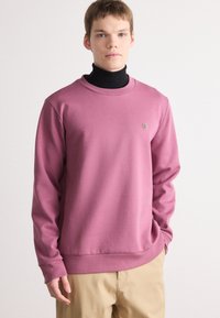 Felpa rosa in tessuto morbido con scollatura rotonda, maniche lunghe e un piccolo logo sul petto, abbinata a pantaloni beige.