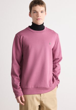 Różowy sweter z miękkiego materiału, z okrągłym dekoltem, długimi rękawami i małym logo na piersi, w zestawieniu z beżowymi spodniami.