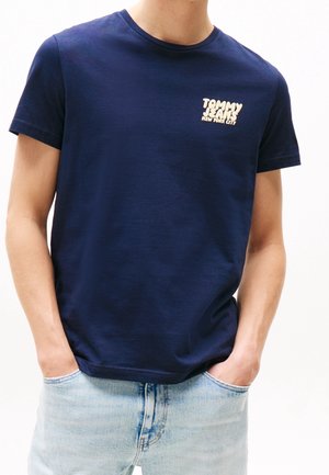 Mężczyzna w granatowej koszulce Tommy Jeans z białym logo na piersi i jasnoniebieskich dżinsach, ręce w kieszeniach na białym tle.