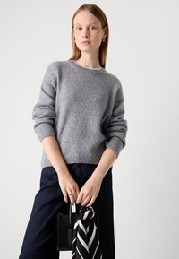 Mulher com suéter cinza e calças escuras segurando uma pequena bolsa preta com um lenço em preto e branco contra um fundo branco liso.