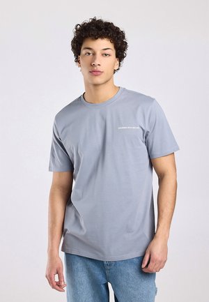 Jonge man met krullend haar, gekleed in een lichtgrijs T-shirt met de tekst "GROWING WITH NATURE" en blauwe jeans, staand tegen een effen achtergrond.