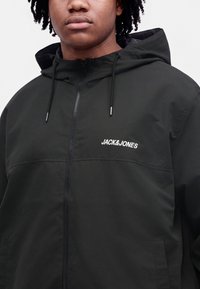 Junge Person trägt eine schwarze Kapuzenjacke mit Frontreißverschluss und dem "JACK&JONES"-Logo auf der Brust.