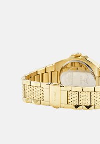 Reloj de pulsera de metal dorado con una correa texturizada, esfera rectangular y corona circular. Presenta un fondo de caja transparente con detalles de marca.