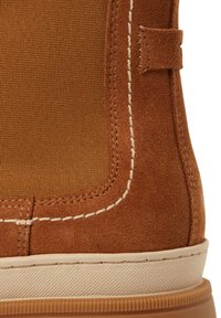 Detailní záběr na hnědou semišovou botu s elastickou postranní částí, bílým kontrastním stehem, poutkem na snadné obouvání a béžovou gumovou podrážkou.