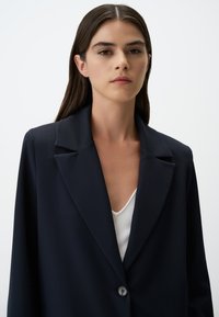 Blazer blu navy con colletto strutturato, tessuto morbido, chiusura con bottone singolo e vestibilità rilassata, indossato sopra un top bianco a coste.