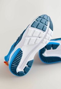 Zapatillas deportivas azules con suelas de goma texturizadas, con una entresuela blanca y un detalle en naranja. Diseñadas para ofrecer agarre y durabilidad, con una forma redondeada.