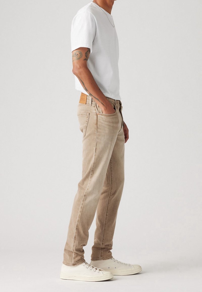 Homme debout de profil, portant un t-shirt blanc, un jean beige, des baskets blanches, avec une main dans la poche et des tatouages visibles sur le bras.