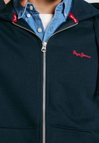 Sudadera con cremallera en navy y cremallera plateada. Presenta un logo rojo en el lado izquierdo del pecho, combinado con una camisa de cuello azul claro debajo.