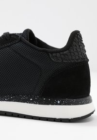 Black athletic shoe featuring a mesh upper, suede accents, textured heel cap, and a speckled midsole with a white base.