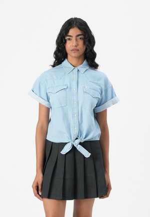 Lys blå denimskjorte med korte oppbrettede ermer, to brystlommer og knyttet foran. Brukt med en plissert svart skjørt.