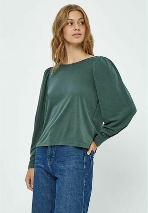 Top long à manches longues vert avec une coupe décontractée, encolure large et manches bouffantes. Tissu doux avec une texture lisse, associé à un jean bleu.