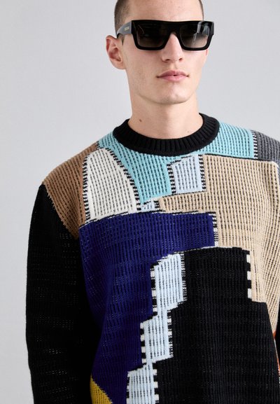 Pull en tricot avec un motif géométrique noir, beige, bleu et violet. Texture côtes avec un col rond noir.