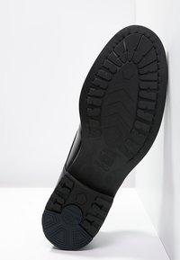 Semelle de chaussure en caoutchouc noir avec un profil texturé, au motif rayé. Elle présente un talon arrondi, des bords lisses et un logo de la marque près de la voûte.