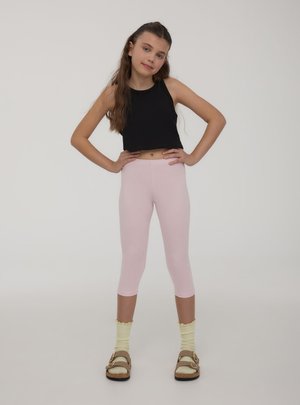 Ragazza con lunghi capelli castani indossa un top corto nero senza maniche, leggings rosa, calzini gialli e sandali marroni, in piedi con le mani sui fianchi.