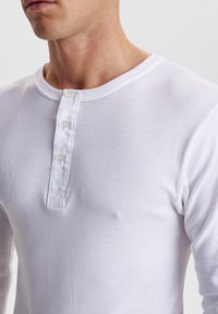 White long-sleeve henley shirt made of soft cotton, featuring a rounded collar and three button placket on the chest. Smooth texture.