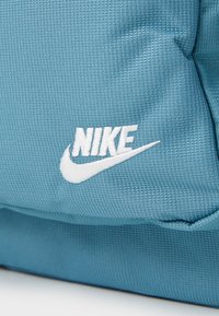 Μπλε υφασμάτο σακίδιο με υφή, που διαθέτει λευκό κεντημένο λογότυπο Nike και swoosh, επιδεικνύοντας έναν ανθεκτικό σχεδιασμό και κλείσιμο με �φερμουάρ.