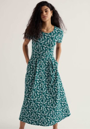 Pikkade lokkis juustega naine, kes kannab sinakashalli lühikeste varrukatega keskmise pikkusega kleiti valge lehemustriga, seisab käed taskutes ühetoonilise tausta ees.