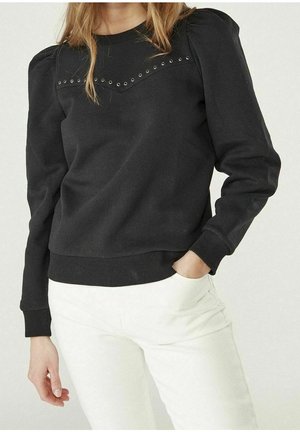 Kobieta w czarnej bluzce z długim rękawem z bufkami na ramionach i metalowymi oczkami, w białych spodniach, z ręką w kieszeni.