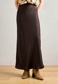 Kvinde iført en mørkebrun satin maxi-nederdel parret med beige ruskind støvletter med hæl på et trægulv.