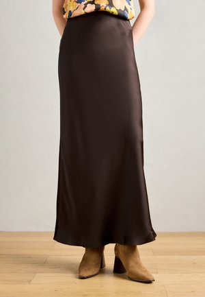 Donna in una lunga gonna maxi di satin marrone scuro abbinata a stivaletti con tacco in suede beige su un pavimento di legno.