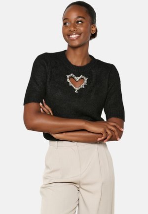 Black knit sweater featuring a heart cutout embellished with floral detailing. Short sleeves and a round neckline. Tailored beige trousers.