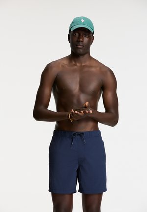 Marineblauwe zwemshorts met een elastische tailleband en koord, gecombineerd met een groene pet met een roze cactusontwerp. Er wordt geen shirt gedragen.