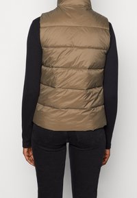 Braune gepolsterte Weste mit hohem Kragen, die über eine horizontale Steppung und eine taillierte Passform verfügt. Getragen über einem schwarzen Langarmshirt.