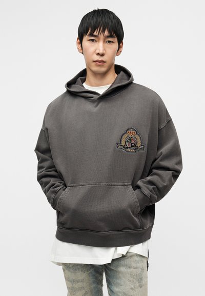 Γκρι φούτερ με κουκούλα από απαλό ύφασμα, με κεντημένο λογότυπο στο αριστερό στήθος, τσέπη καγκουρό και άνετη εφαρμογή. Φορεμένο πάνω από λευκό πουκάμισο.