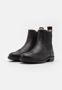 Botas de cuero negro hasta el tobillo con detalles de brogue, paneles laterales elásticos y una suela texturizada. Forro suave visible en la abertura.