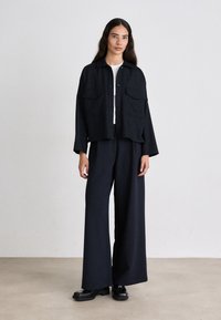 Schwarze, gestreifte Cropped-Jacke mit zwei vorderen Taschen, kombiniert mit weit geschnittenen Hosen. Aus glattem Stoff und glänzenden schwarzen Schuhen.