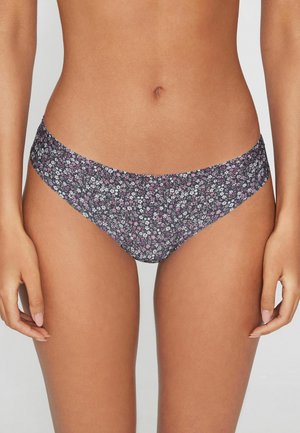 Žena nosící bikiny s nízkým pasem s malým růžovým a bílým květinovým vzorem na tmavém pozadí proti šedému pozadí.