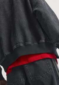 Črna jeans jakna z rebrastim robom in manšetami, oblečena čez rdečo srajco, ki razkriva rob srajce. Ima sproščen kroj in teksturirano tkanino.