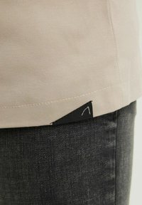 Beige tygkantsöm med synlig söm och en liten svart triangulär etikett ovanför mörkgrå denimjeans.