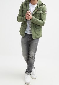 Grøn bomuldsjakke med flere lommer og hætte, båret over en hvid skjorte, parret med grå jeans og hvide sneakers.