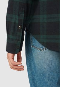 Ciemnozielona i czarna koszula flanelowa w kratę z mankietami z czarnymi zatrzaskami, noszona na jasnoniebieskich dżinsach z detalem w postaci tylnej kieszeni.