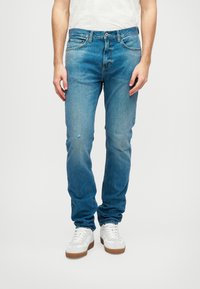 Μπλε τζιν παντελόνι με ευθεία γραμμή, που διαθέτει ξεθωριασμένο φινίρισμα, ελαφρές φθορές και πέντε τσέπες, συνδυασμένο με λευκά αθλητικά παπούτσια.